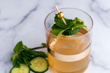 A light amber cocktail served in a short glass with a large ice cube, garnished with a cucumber slice and fresh mint leaves skewered on a decorative pick.