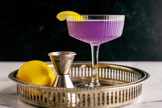 A vibrant purple cocktail in a coupe glass garnished with a lemon twist, displayed on a silver tray with a fresh lemon and jigger—ideal for a craft cocktail blog or gin-based drink recipe.