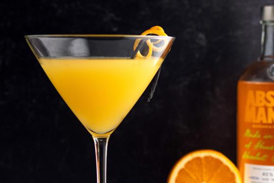 Bright orange martini cocktail in a stemmed glass, garnished with a vanilla bean and orange twist, with Absolut Mandarin vodka bottle and fresh orange slice in background.