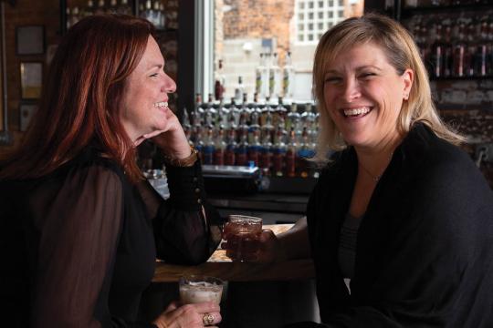 Karen and Jen sit together at a bar. Karen faces the camera with a smile, while Jen, also smiling, turns toward Karen.