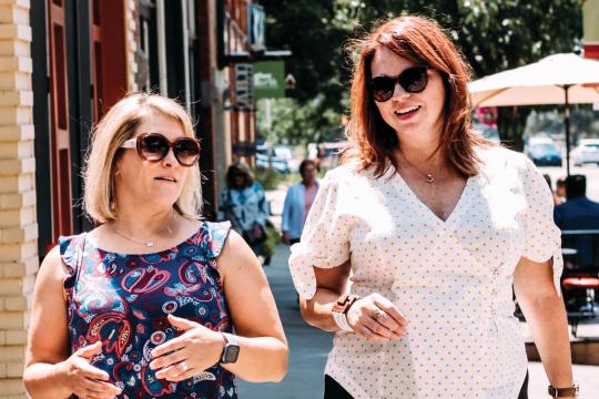 Karen (left) and Jen (right) walk and talk on a sunny downtown sidewalk, both wearing sunglasses and summer tops, engaged in a casual, animated conversation.