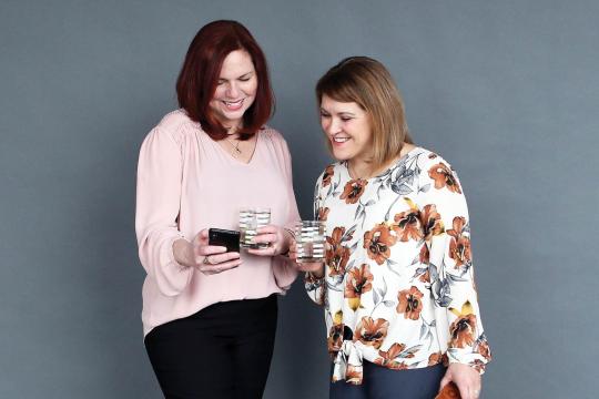 Jen and Karen, cohosts of The Speakeasy Podcast, laughing together while looking at a phone and holding cocktails. Jen wears a blush blouse and Karen wears a floral top with orange and gray flowers.