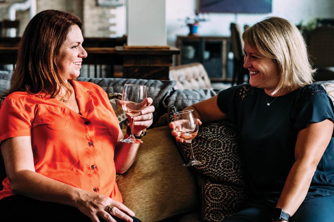 Jen, owner of Redhead Creative Consultancy, and Karen, cohosts of The Speakeasy Podcast, sitting on a sofa and laughing while holding glasses of rosé wine.