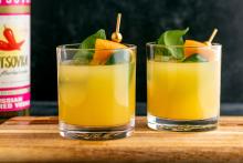 Two glasses of an orange drink garnished with a basil leaf, orange peel, and jalapeno. 