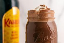 A close-up of a glass mason jar filled with frothy, rich hot chocolate, topped with whipped cream and a dusting of cocoa powder. In the blurred background, a bottle of Kahlúa, a coffee-flavored liqueur, is partially visible, indicating that the drink contains the liqueur.