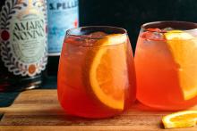 Refreshing summer cocktail with orange slices and ice, featuring Amaro Nonino and sparkling water, served in stemless glasses on a wooden board for The Speak Easy Podcast.