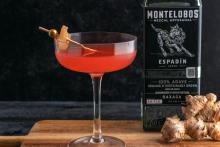 Red cocktail with orange garnish next to Montelobos Espadín mezcal bottle on wooden surface, craft cocktail photography for Speak Easy Podcast.