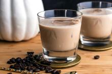 A creamy iced coffee cocktail for the Speak Easy podcast served in a short glass over ice, sitting on a wooden surface with scattered coffee beans and a gold bar spoon nearby.