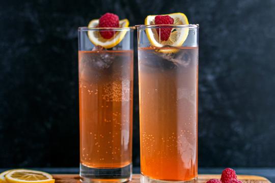 Tall cylinder glasses filled with an amber liquid. The glasses are garnished with a raspberry and lemon slice.