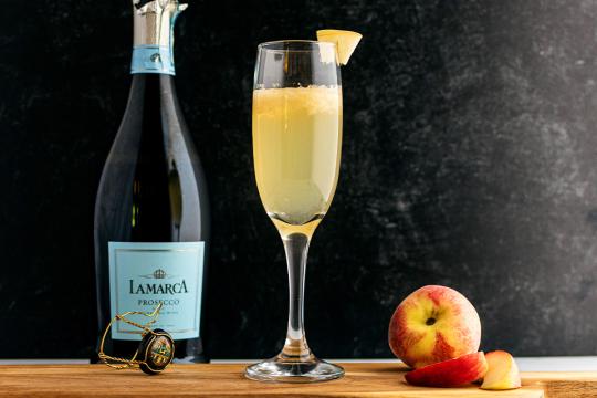 From left to right, the image features a bottle of prosecco, a wine glass fill with a pale yellow liquid, and a peach.