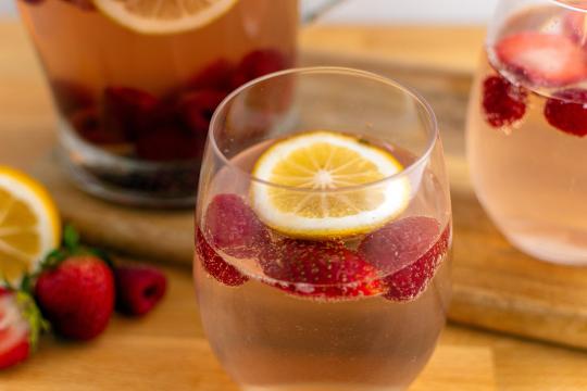 Photo of a wine glass filled with light pink liquid, with strawberry slices and a lemon slice floating on top.