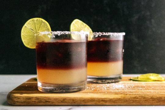 Two short cocktail glasses, filled with an amber liquid with a clear line of dark red liquid resting on top.