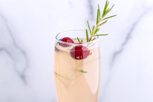 A champagne flute filled with a pale peach liquid, garnished with two cranberries and a sprig of rosemary.