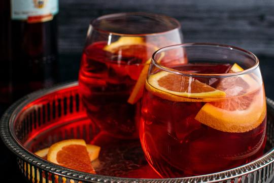 Two short round glasses filled with a red cocktail, garnished with orange slices and served over ice, sit on a silver tray.