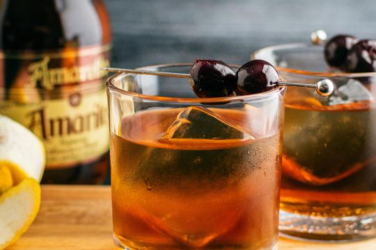 Bourbon cocktail served over a large ice cube, garnished with cocktail cherries. Classic amaretto drink with bottle and lemon peel in background.