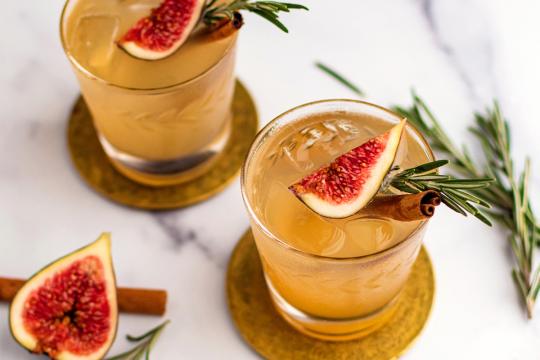 Golden cocktails with fresh fig, rosemary, and cinnamon garnish on brass coasters, fall cocktail photography for Speak Easy Podcast.