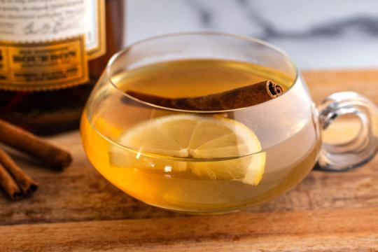 Hot toddy cocktail with lemon slice and cinnamon stick in glass teacup with bourbon bottle, warm winter drink photography for Speak Easy Podcast.