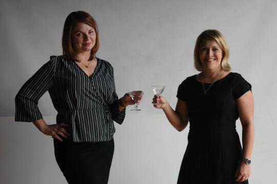 Jen and Karen standing against a grey backdrop, holding cocktail glasses