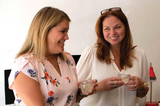 Karen and Jen are standing side by side, holding small cocktail glasses. It looks like they are about to start laughing.