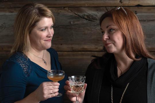 Karen (left) and Jen (right) clink glasses and share a smile while holding cocktails, standing in front of a rustic wooden wall.