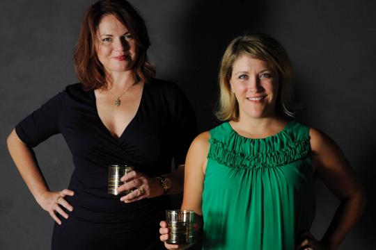 Hosts of The Speakeasy Podcast, Jen and Karen, holding cocktails and smiling in front of a dark backdrop. Jen wears a black dress and Karen wears a green top.