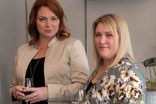 Jen and Karen, co-hosts of Speak Easy Podcast, professional portrait with wine glasses in modern kitchen setting.