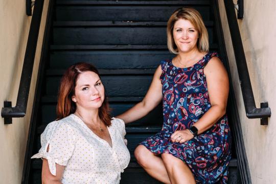 Jen and Karen, cohosts of The Speakeasy Podcast, sitting together on black stairs. Jen wears a white blouse with gold dots, and Karen wears a navy paisley dress.