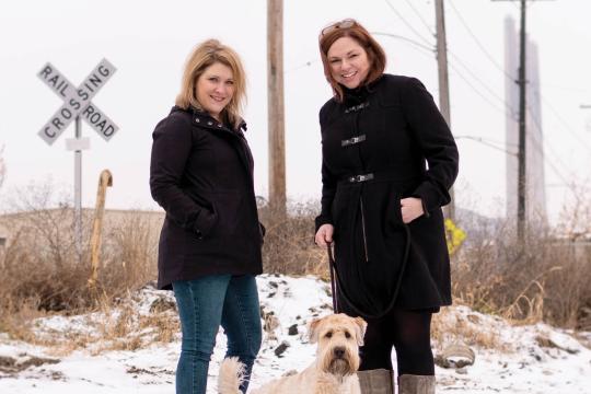Karen and Jen, co-hosts of Speak Easy Podcast, with dog at railroad crossing in winter outdoor lifestyle photo.