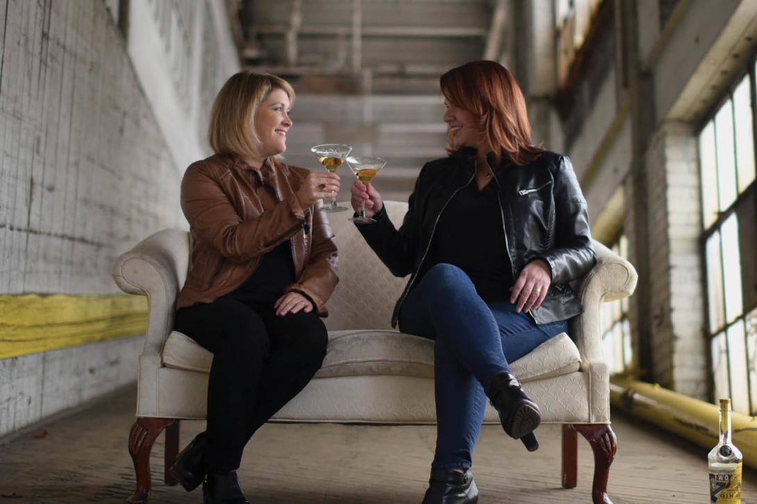 Karen and Jen are sitting side by side on a couch. Their glasses are raised in a toast.