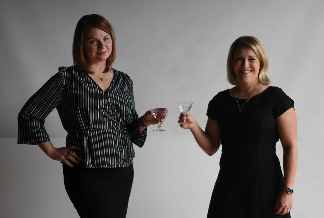 Jen and Karen standing against a grey backdrop, holding cocktail glasses