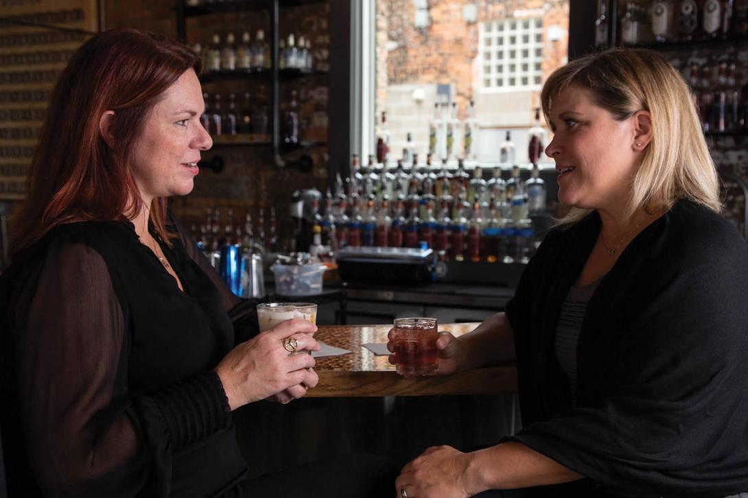 Jen and Karen are sitting side by side at a bar, facing each other with drinks in their hands.