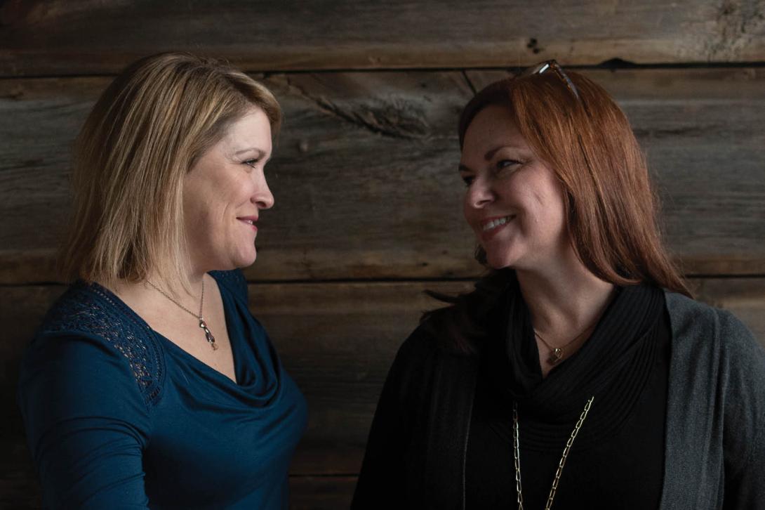 Picture of Karen and Jen smiling towards each other, with a brick wall background.