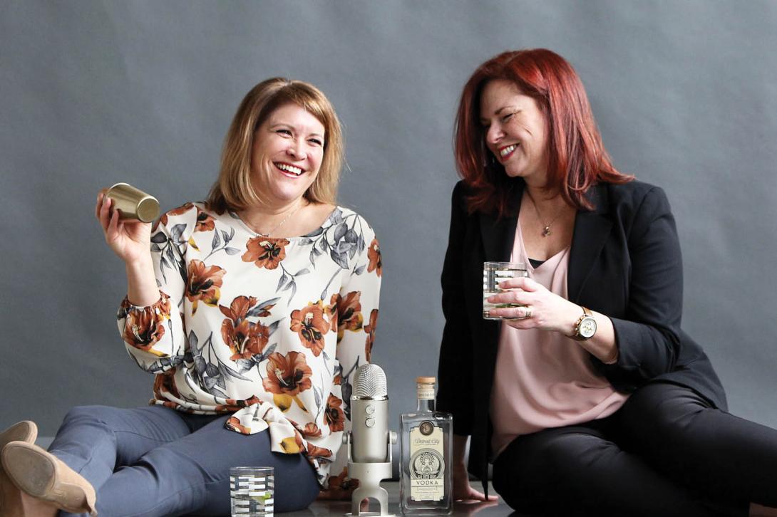 Picture of Karen and Jen sitting on the floor next to each other. Karen is holding a cocktail shaker, and between them is a podcast microphone.