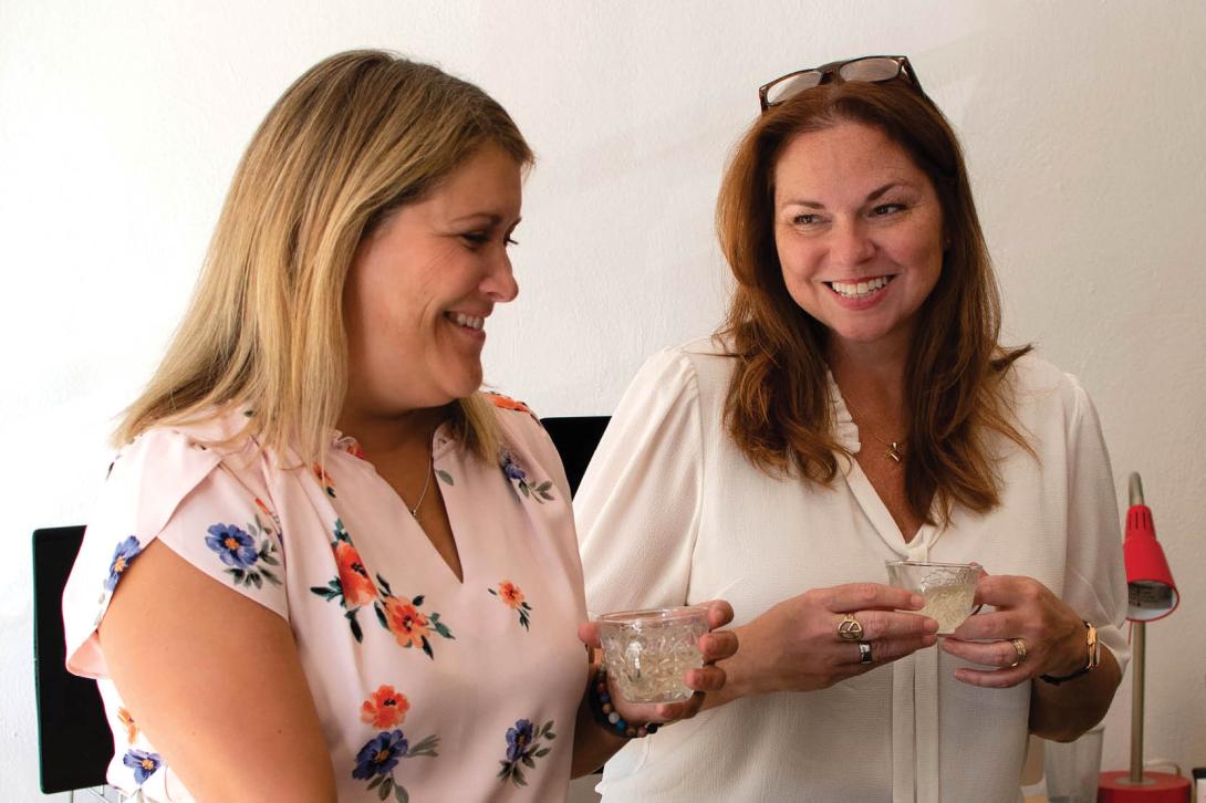 Karen and Jen are standing side by side, holding small cocktail glasses. It looks like they are about to start laughing.