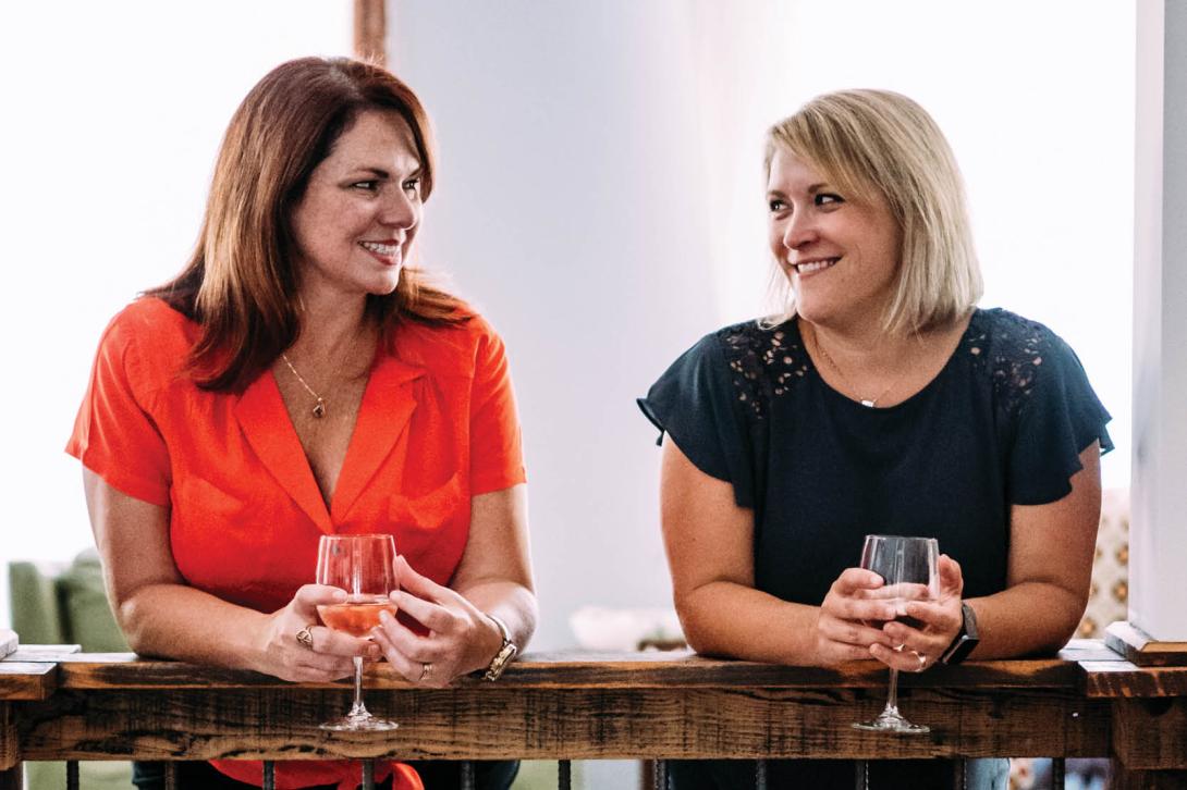 Jen and Karen, co-hosts of The Speakeasy Podcast, chatting over wine at a cozy indoor location. Jen wears a red blouse; Karen wears navy.